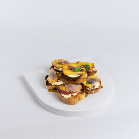 Arch quartz serving platter in Caesarstone Alpine Mist created by Aureliia Collection. Similar to a marble platter, the Arch Quartz platter shown with peach and prosciutto crostini. An excellent home decor gift for someone who has it all.