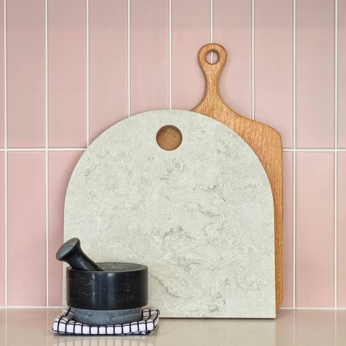 Large arch cheese platter with timber paddle board and black pestle and mortar. Pastel pink tiled splashback. Caesarstone Bianco Drift.